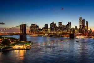 Elevated view of the Brooklyn Bridge and Lower Manhattan skyscrapers at dusk. The illuminated skyline of the Financial District reflects in the East River. New York City.
