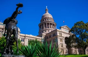 Cavalcando un cavallo a sinistra del Texas State Capital edificio in una bella giornata cielo blu chiaro con la luce del sole colpire la parte anteriore del capitol piante verdi fresche in primo piano con il governo