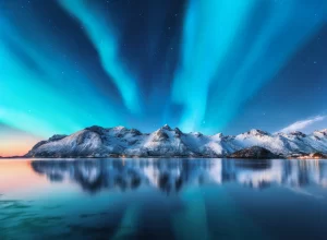 Aurora boreale e montagne innevate nelle isole Lofoten, Norvegia. Aurora boreale. Cielo stellato con luci polari e rocce innevate riflesse nell'acqua. Paesaggio invernale notturno con aurora, mare.
