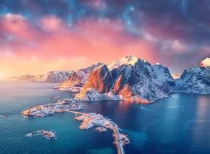 Beautiful landscape with blue sea, snowy mountains, rocks and islands, village, rorbu, road, bridge and pink sky at sunrise. Aerial view. Hamnoy in snow in winter in Lofoten islands, Norway.