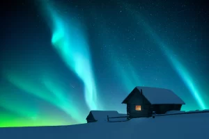 Fantastic winter landscape with wooden house with light in window in snowy mountains and northen light in night sky. Christmas holiday and winter vacations concept