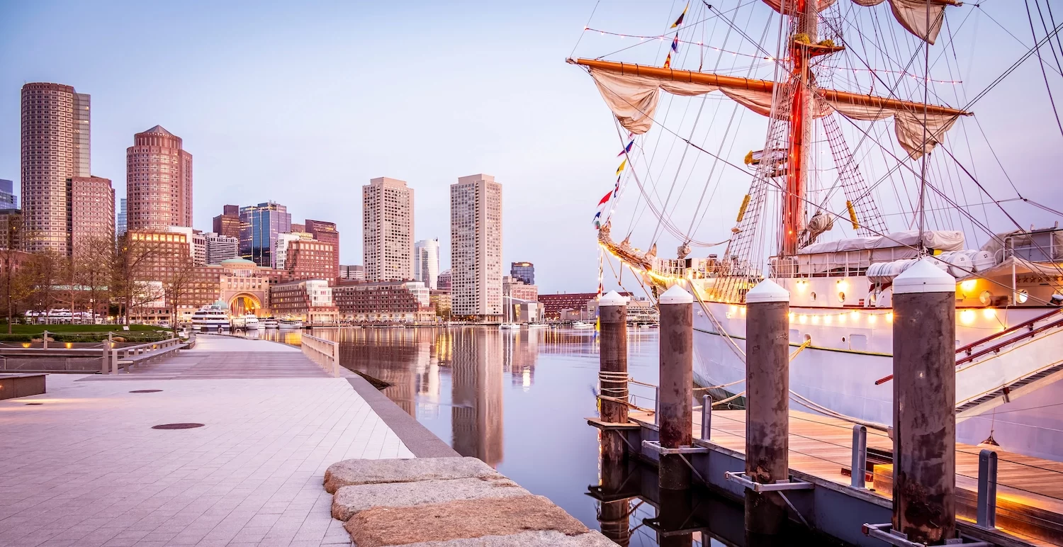 The historic architecture of Boston in Massachusetts at sunrise at Fan Pier with the iconic skyline of the city and a vintage boat anchored on the side with lights on.