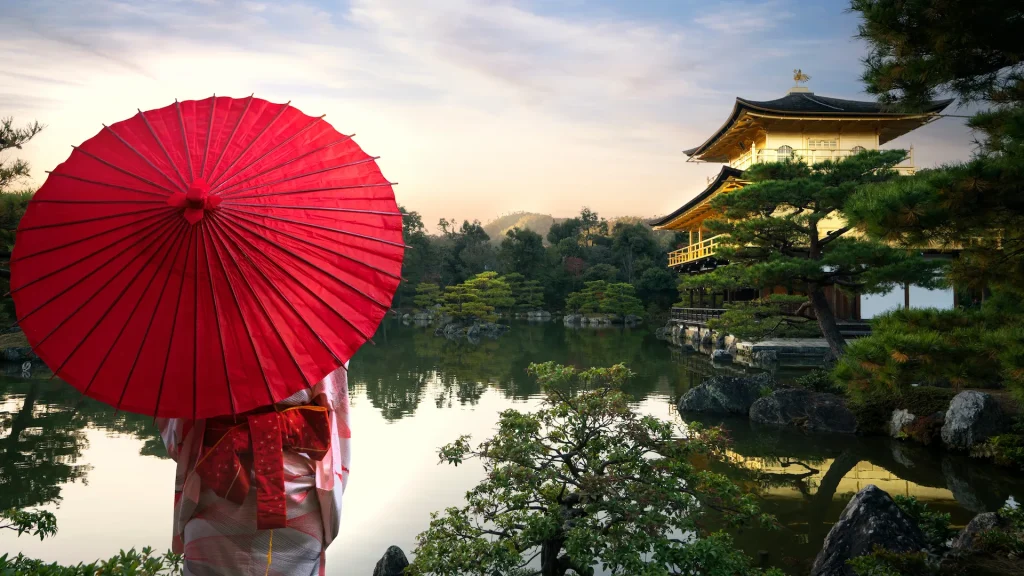 Viaggiatrice asiatica in kimono, abito tradizionale giapponese, con un ombrello rosso, in piedi nel tempio Kinkakuji dorato in un parco autunnale nella città di Kyoto, Giappone | Tour combinato Giappone e Thailandia | Alidays Travel Experience