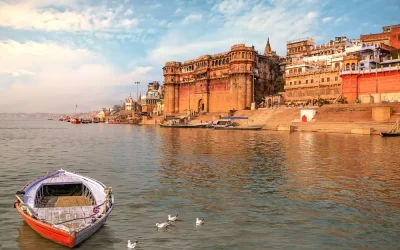 Ancient Varanasi India city architecture with view of Ganges river ghat at sunset. Wooden boat seen on river Ganga with migratory birds