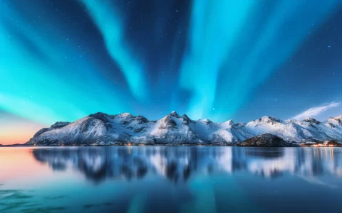 Aurora boreale e montagne innevate nelle isole Lofoten, Norvegia. Aurora boreale. Cielo stellato con luci polari e rocce innevate riflesse nell'acqua. Paesaggio invernale notturno con aurora, mare.